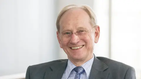 Renishaw Sir David McMurtry pictured smiling at the camera. He has short, light-coloured hair and wears round frameless glasses. He also wears a light blue shirt and a darker blue tie with a diamond pattern on it, and a grey suit jacket. 