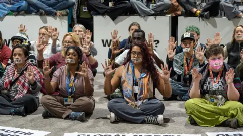 BBC Climate activists sitting on the floor wearing tape over their mouths, with "Pay Up" written on the tape. 