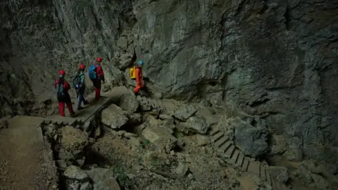 Xiqing Wang/ BBC Four people climb down rock stairs into the hole