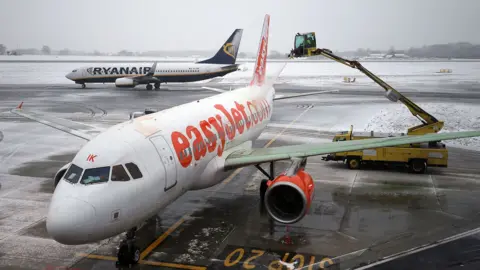 PA Media An Easyjet branded aeroplane is de-iced by an automated crane-style cart during cold weather, with a Ryanair branded aeroplane on the runway in the background, and with snow and ice all around, at Luton airport in 2017.