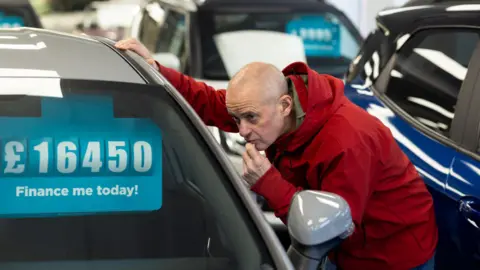Getty Images Man in red hooded jacket looks in a car window at a vehicle in a showroom