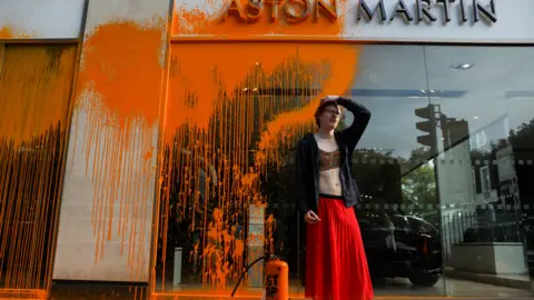 AFP A Just Stop Oil activist stands in front of an Aston Martin showroom in central London, with orange paint splattered across the glass facade. The activist, wearing a red skirt and black jacket, holds a paint sprayer labeled "JUST STOP OIL" at their feet.