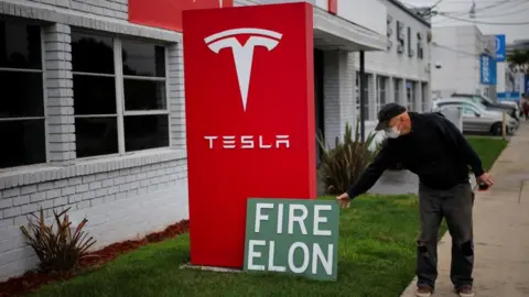 Reuters A man places a sign saying ''Fire Elon'' outside a Tesla dealership during a protest against Tesla and CEO Elon Musk in Los Angeles