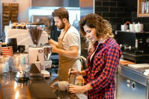 Getty Images A young woman with long wavy hair and a red checked shirt is decorating a cup of cappuccino. A young bearded male barista, wearing a beige apron and grey t-shirt, is working in the background amidst steam rising from an espresso machine. The café has a sleek black-tiled wall and modern coffee-making equipment, creating a warm and inviting workspace atmosphere.
