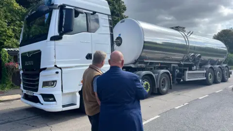 Jonah Fisher/BBC A water tanker drives past two men who watch by the side of the road. 