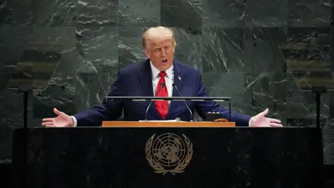 Getty Images President Trump, in a navy blue suit, white shirt and red tie, stands behind a UN lectern giving a speech. He has both arms outstretched.
