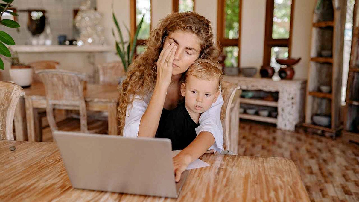 An exhausted mother uses her laptop while her son sits on her lap.