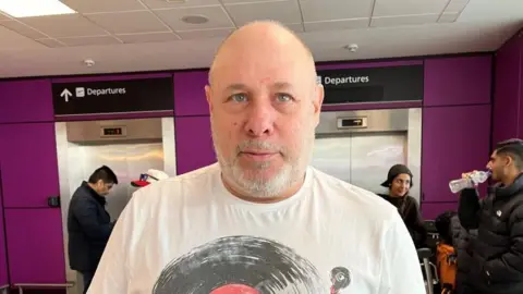 Jeff is bald with a grey beard. He has blue eyes. He has a white t-shirt on. The airport is in the background.