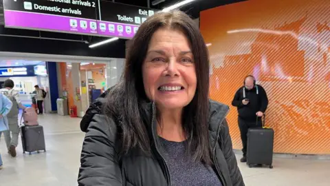 Christine is smiling at the camera. She has long dark hair and is wearing a black coat and top. The airport is in the background.
