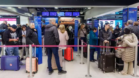 EPA Passengers standing in line with luggage at a British Airways check-in area inside an airport terminal, with digital flight information screens visible above the counters.